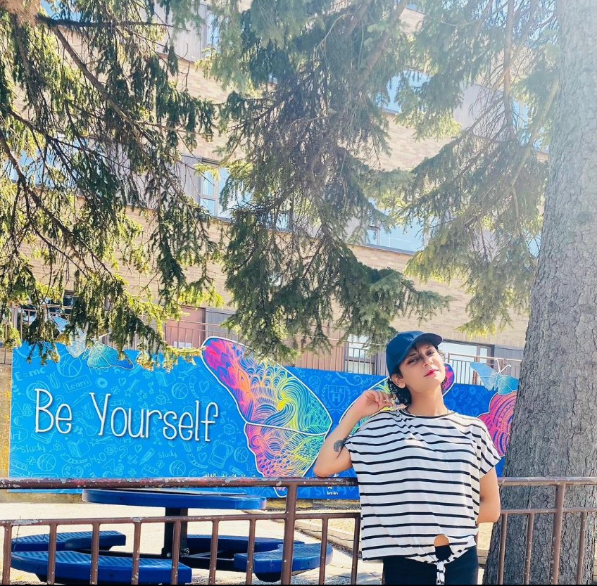 A photograph of Farida in front of a mural that reads "Be Yourself."
